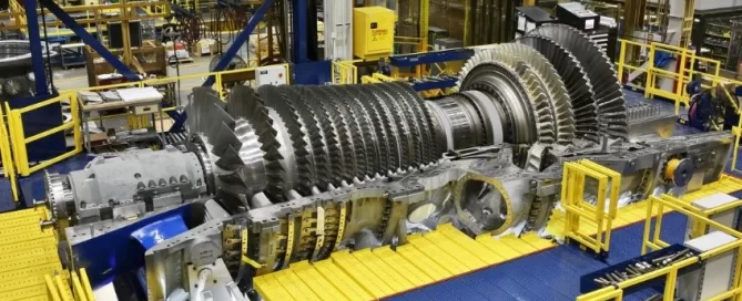A large industrial turbine assembly sits in a manufacturing facility, surrounded by yellow and blue safety barriers, with machinery and equipment visible in the background.