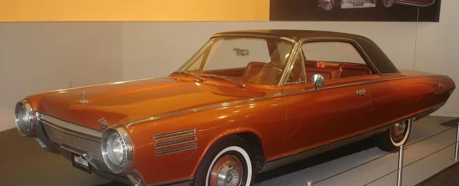 A vintage bronze car with a sleek, aerodynamic design is on display in a showroom, featuring a hardtop roof and circular headlights.