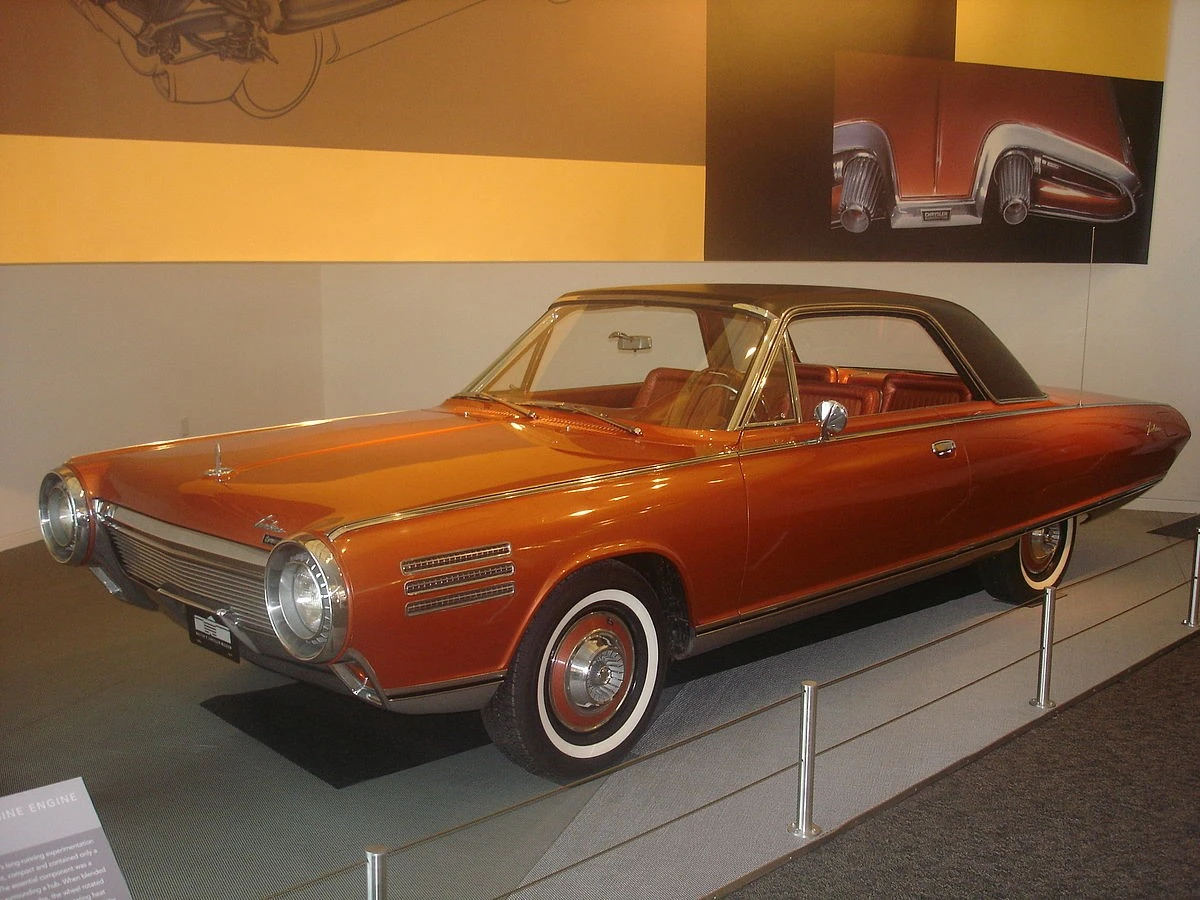 A vintage bronze car with a sleek, aerodynamic design is on display in a showroom, featuring a hardtop roof and circular headlights.