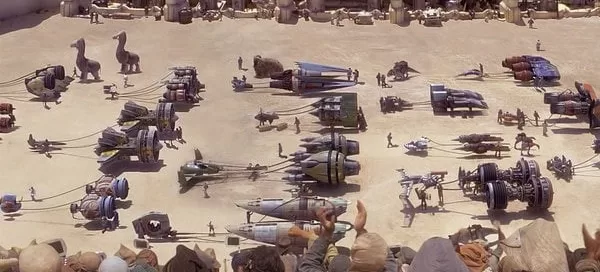 A desert arena with an audience overlooks various futuristic racing pods and vehicles, along with droids and people preparing for a race.