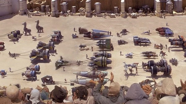 A desert arena with an audience overlooks various futuristic racing pods and vehicles, along with droids and people preparing for a race.