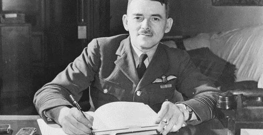 A man in a military uniform sits at a desk, writing in a notebook.