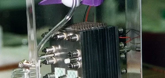 A transparent box encloses a mechanical device from Softinway, with metal components and wires, positioned on a wooden surface. A small purple turbomachinery fan is mounted on top of the apparatus.