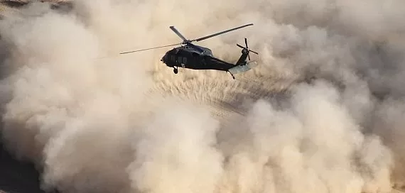 A helicopter hovers above a sandy desert, creating a large dust cloud.
