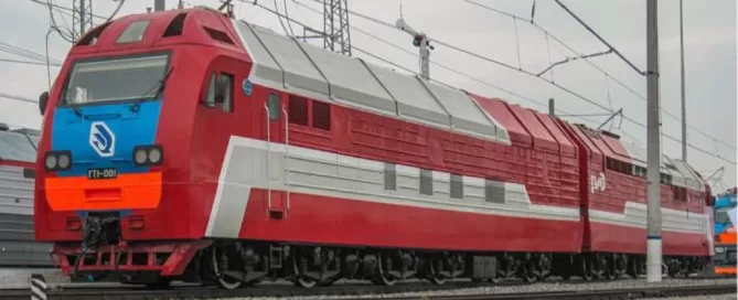 The red and gray train with multiple sections stands majestically on the tracks, resembling a marvel of turbomachinery, surrounded by a network of overhead power lines.