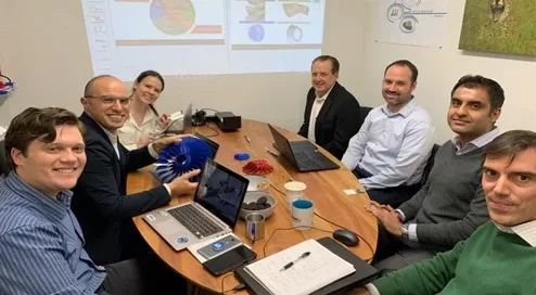 Seven people seated around a table having a business meeting with laptops and documents, discussing turbomachinery solutions. A Softinway presentation is on the screen, accompanied by wall posters in the background.