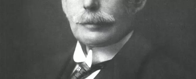 Black and white portrait of an older man with glasses, a mustache, and receding hairline wearing a suit and tie. Beige background.